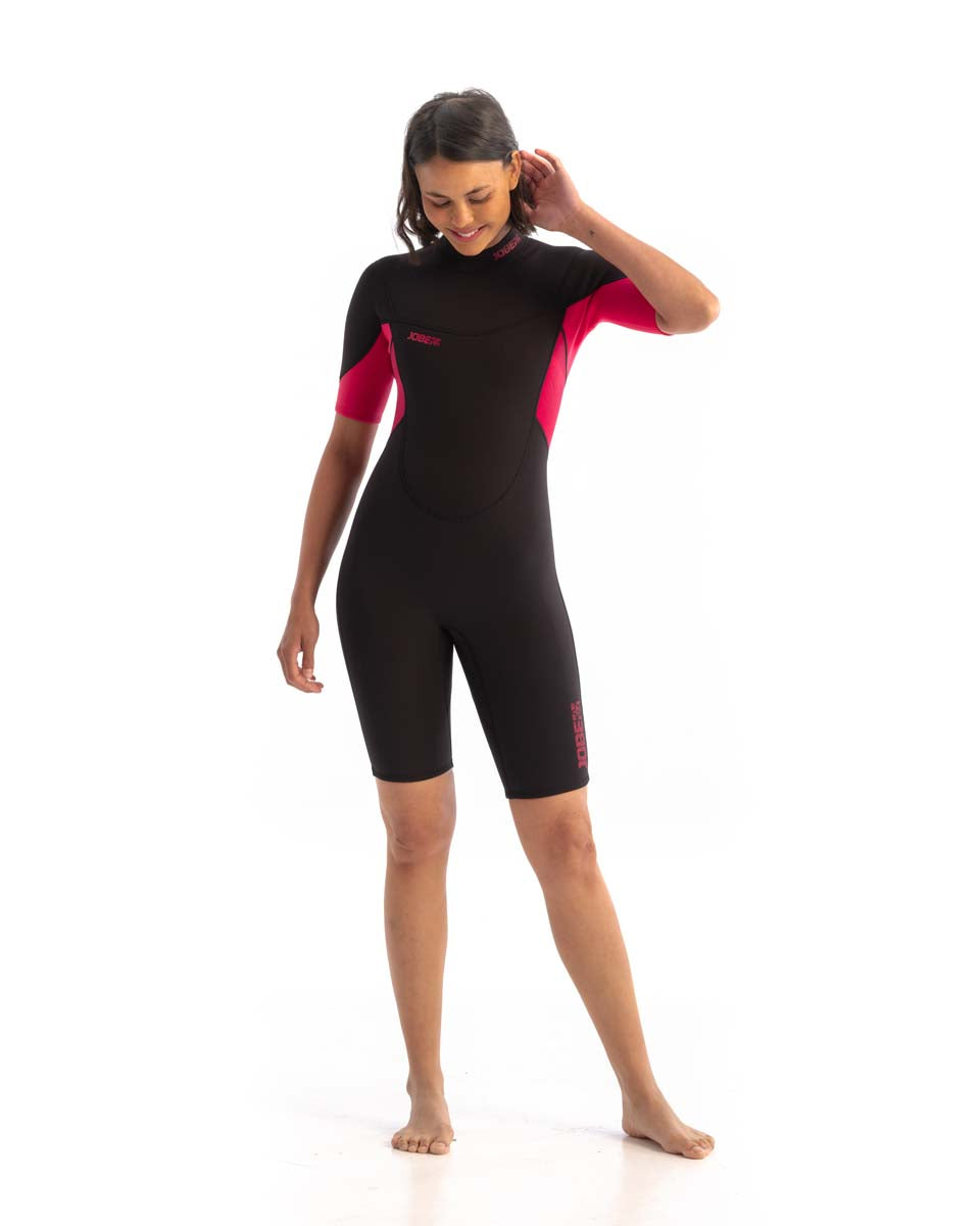 A woman standing and smiling, wearing a black and hot pink Jobe womens shorty wetsuit with short sleeves and short legs.
