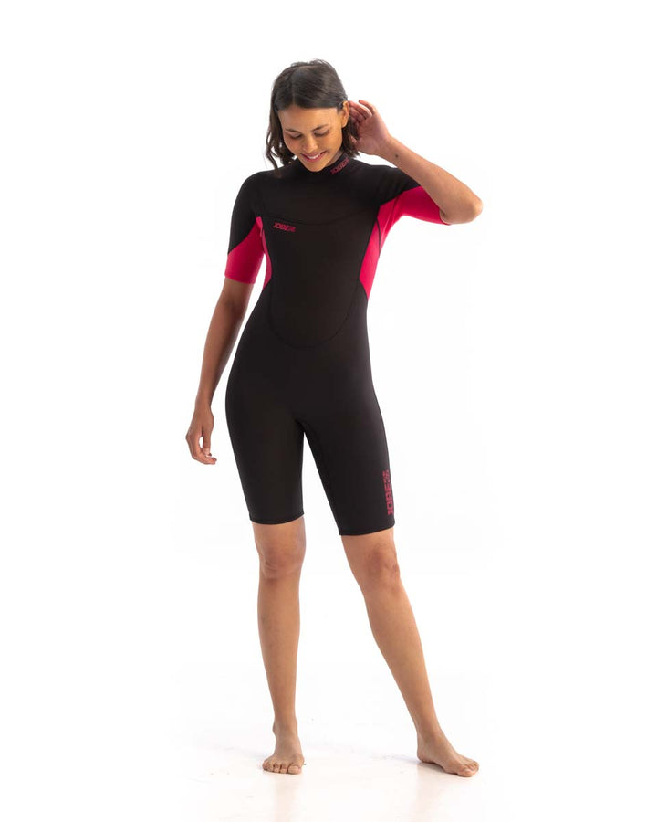 A woman standing and smiling, wearing a black and hot pink Jobe womens shorty wetsuit with short sleeves and short legs.