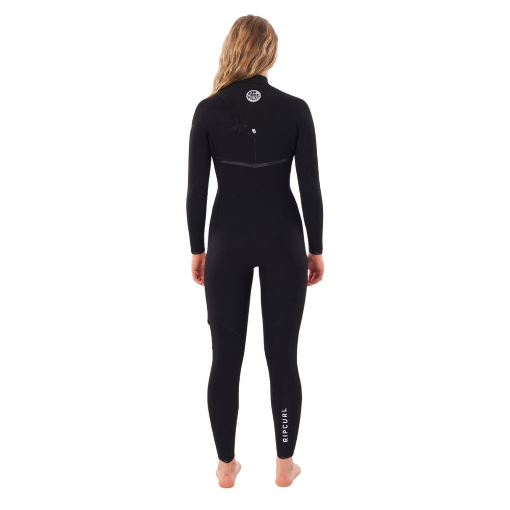 A woman standing and facing forward, wearing a black full-body e bomb zip free womens wetsuit with the Rip Curl brand logo on the chest. View of the back