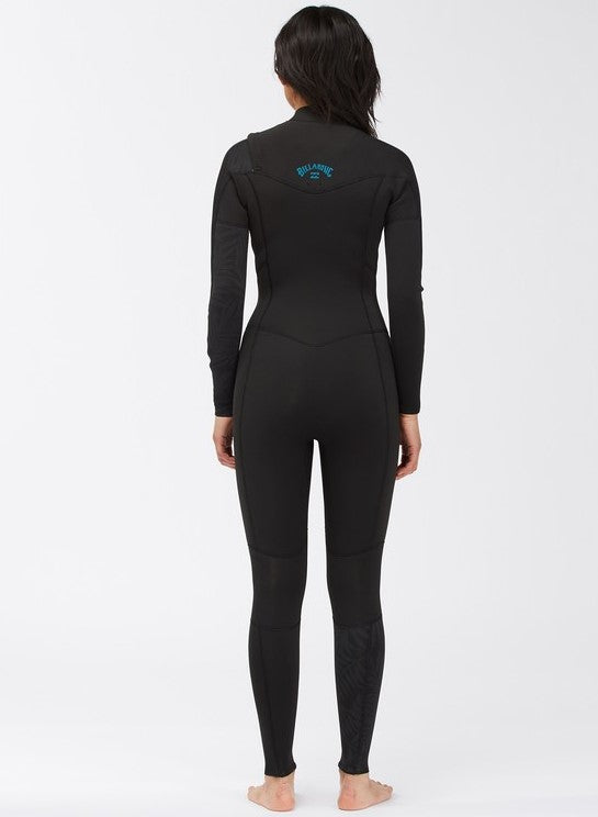 A woman standing straight with her back facing forward, wearing a Billabong wetsuit. The wetsuit is black with the brand logo visible on the upper back area.