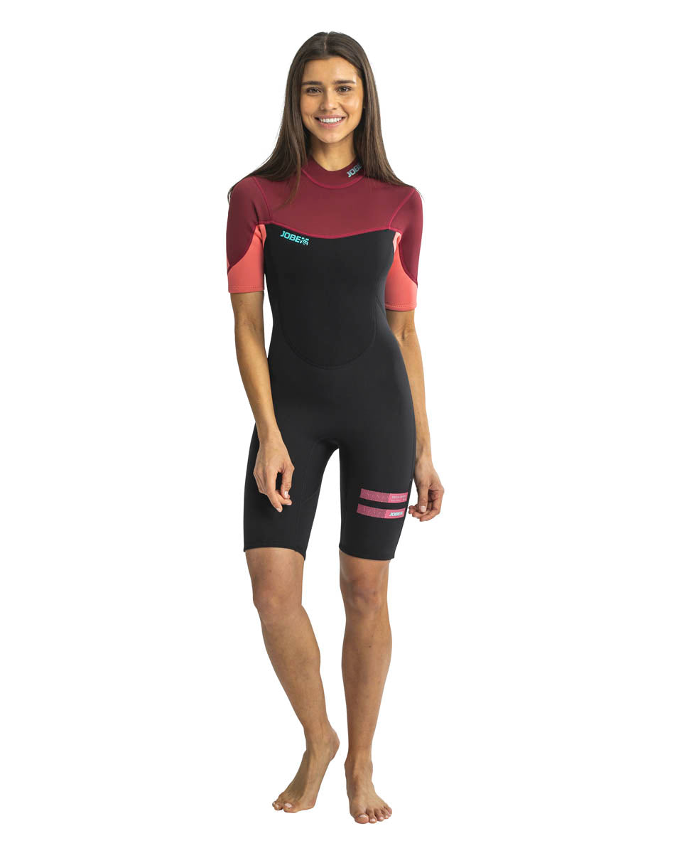 A woman standing and smiling, wearing a red and black Jobe womens shorty wetsuit with short sleeves and short legs.