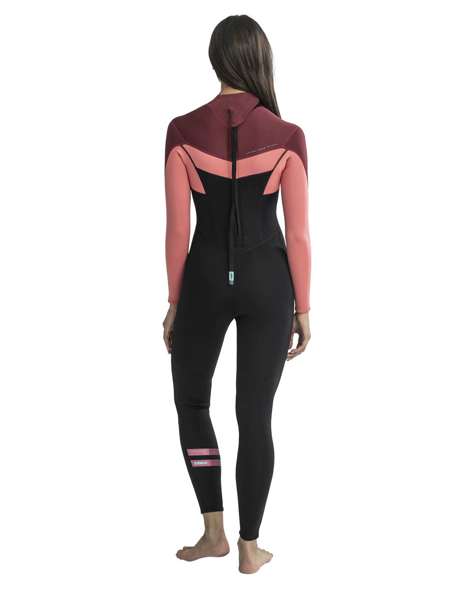 A woman standing, wearing a black and red, and rose pink Jobe wetsuit with long sleeves and a back full zipper. Back view.