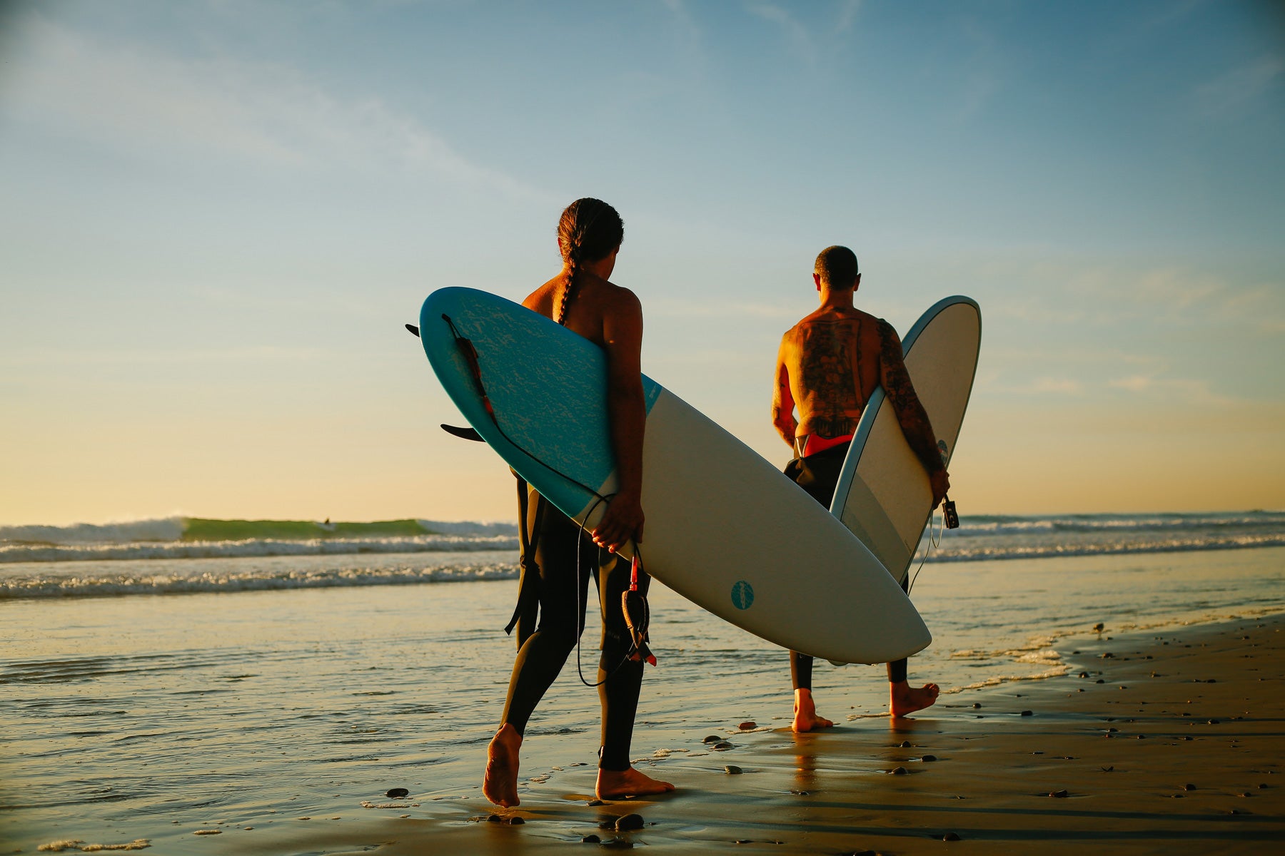 2 Surfers walking on beach carrying surfboards and checking out the waves. 