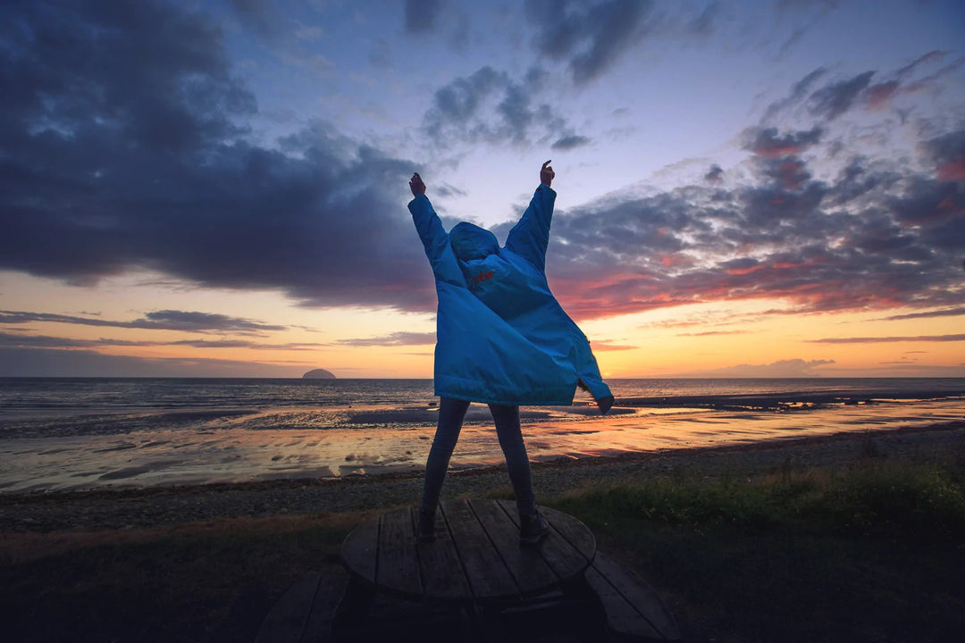 Person on the beach at susnet with their arms up in the air while wearing a blue Dryrobe changing robe. 