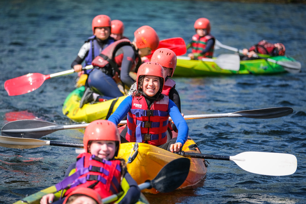 Children kayaking in a body of water with life vests and helmets.