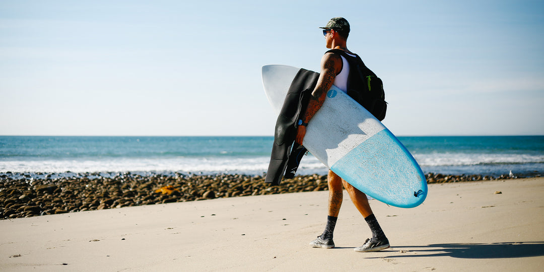 Man walking to the beach carrying his NSP surfboard and wetsuit. 