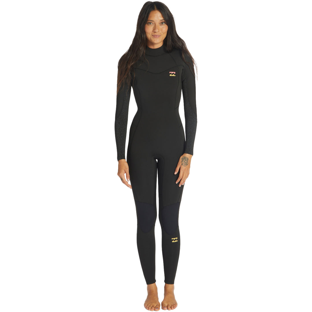 A woman standing and posing in a black full-body Billabong Synery 5/4 womens wetsuit with a back zip entry.