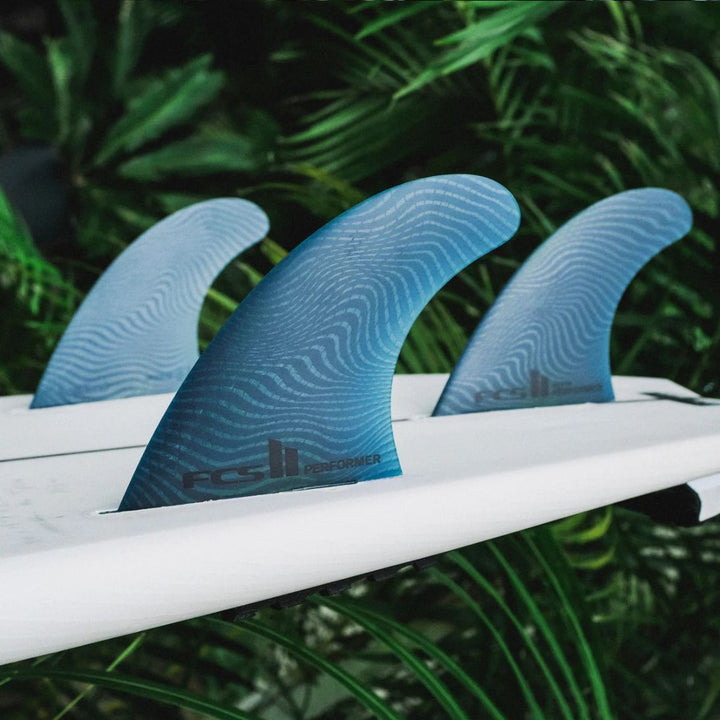 Three blue surfboard fins on a white surfboard with a green leafy background