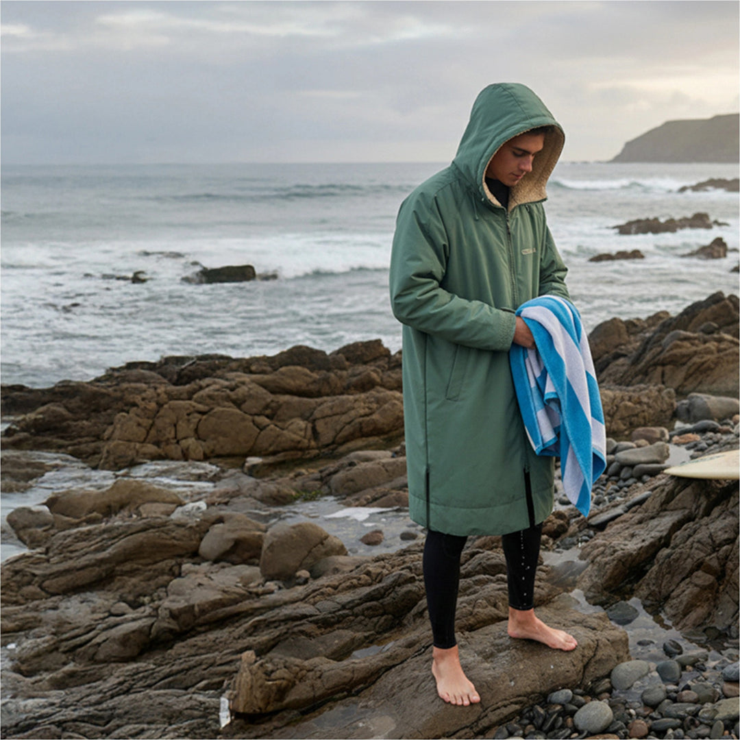 Surfer drying off in a green Gul Revorobe on a rocky beach