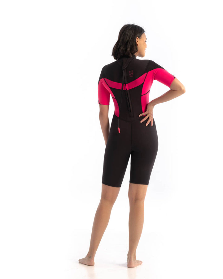 A woman standing, wearing a  black and hot pink Jobe womens shorty wetsuit with short sleeves and short legs. View of the back.