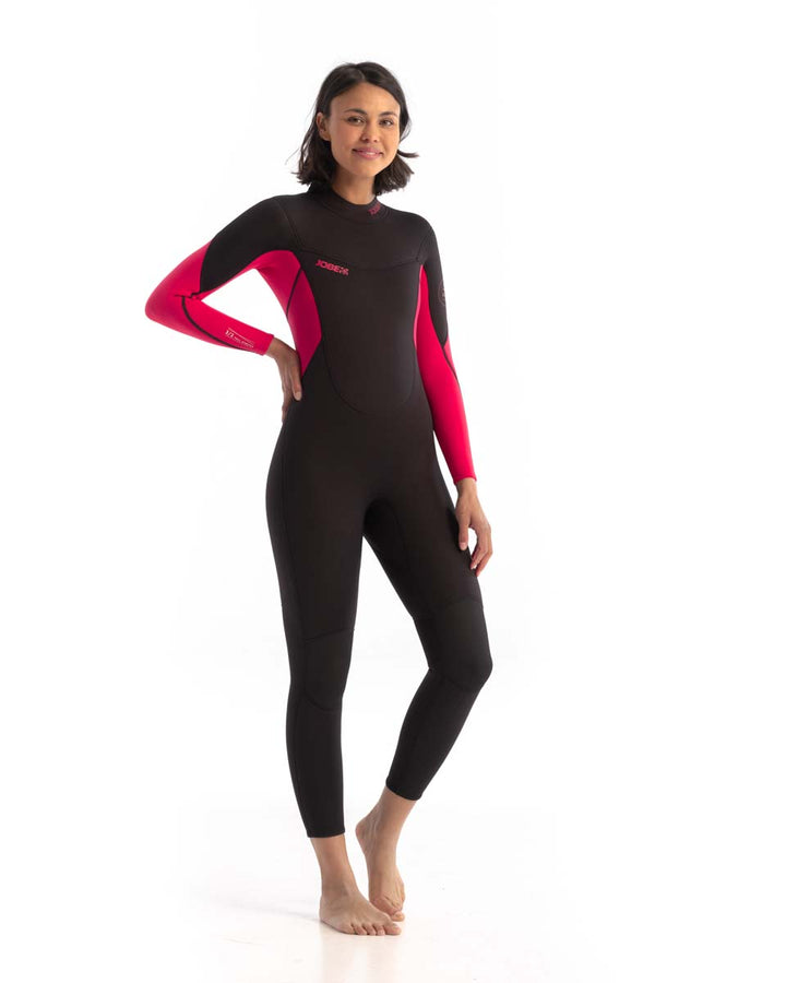 A woman standing, wearing a black and pink Jobe Sofia wetsuit with long sleeves and a back full zipper. Front view