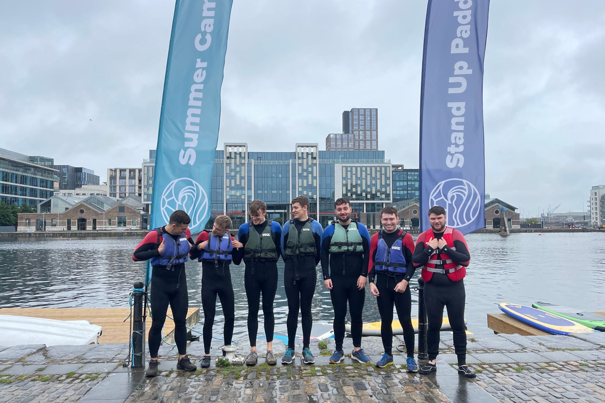 Group of people in wetsuits standing by a body of water with 'Stand Up Paddle' banners in the background.