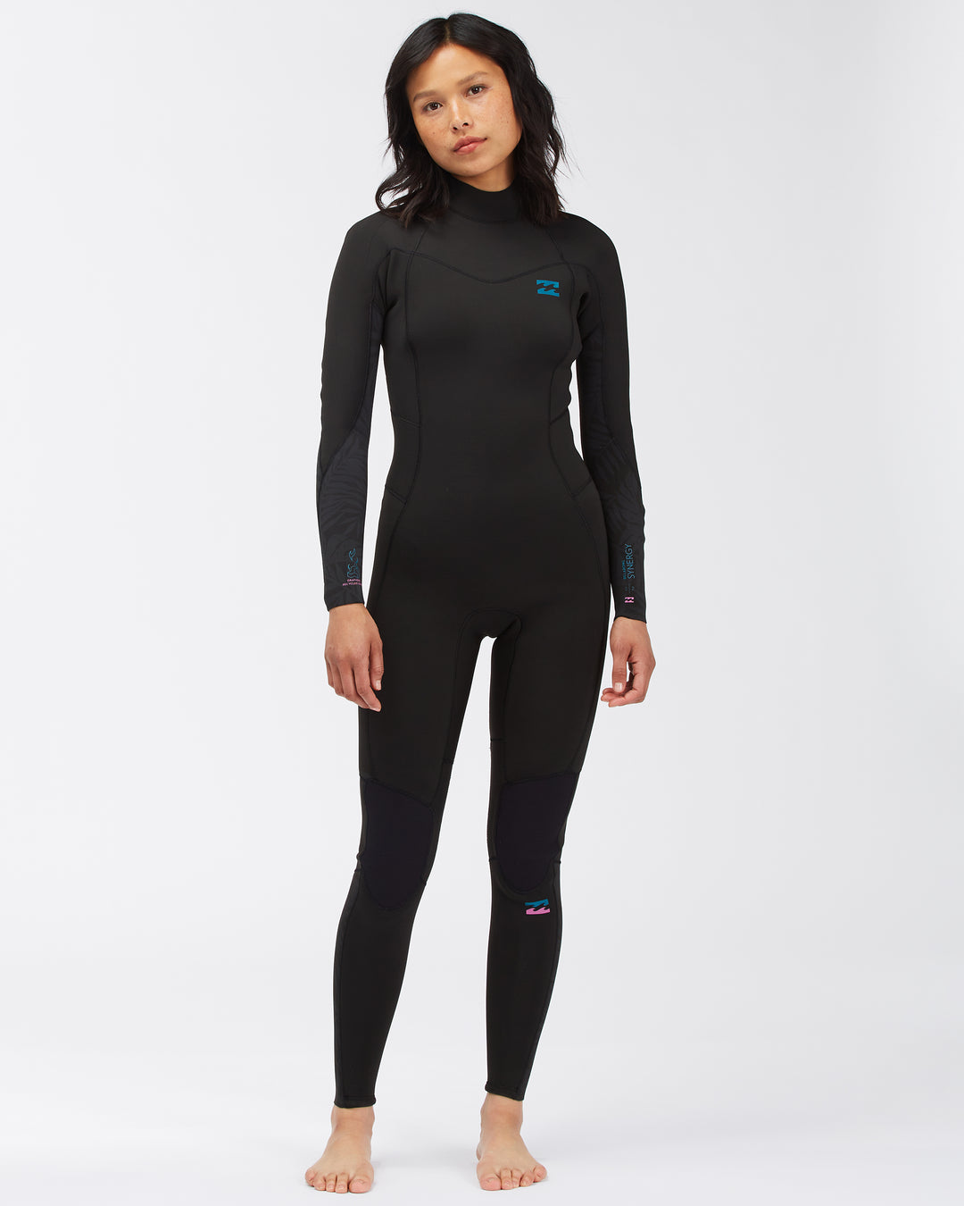 A woman standing and posing in a black full-body Billabong Synery 5/4 womens wetsuit with a back zip entry.