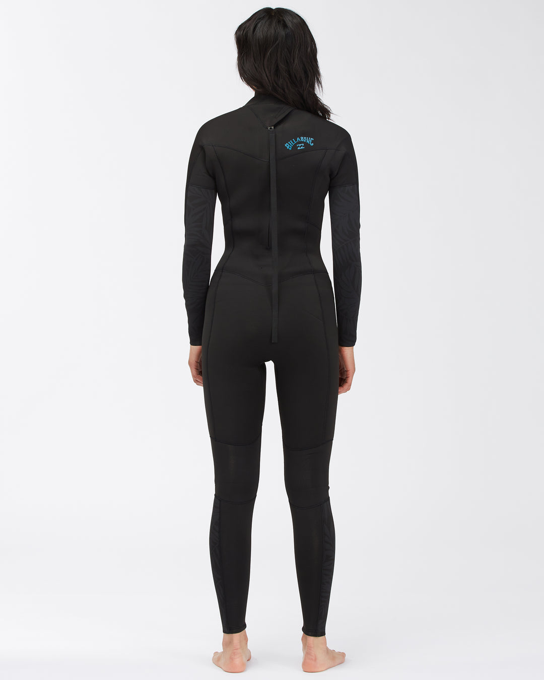 A woman standing with her back facing and posing in a black full-body Billabong Synery 5/4 womens wetsuit with a back zip entry. 