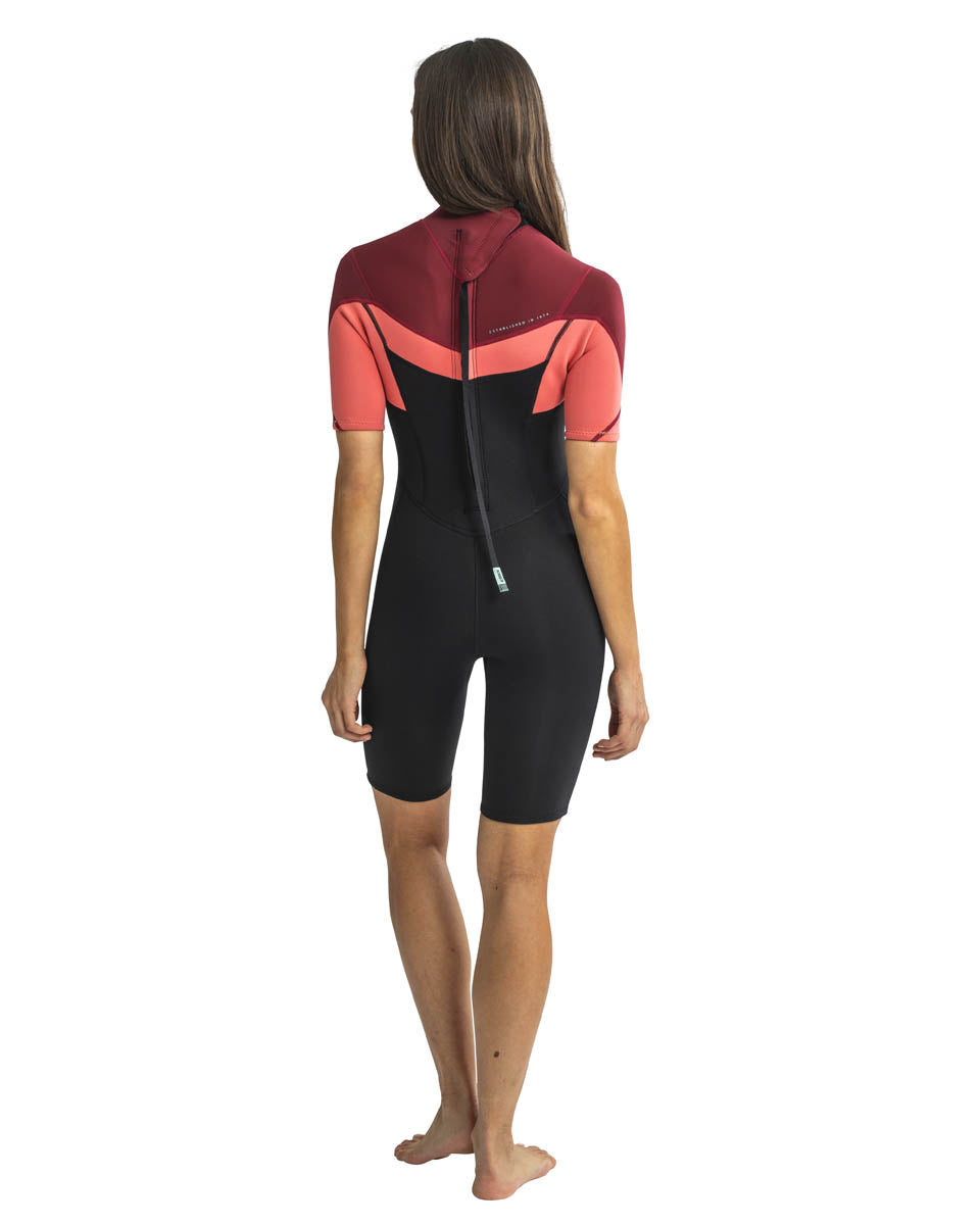 A woman standing, wearing a red, pink and black Jobe womens shorty wetsuit with short sleeves and short legs. View of the back. 