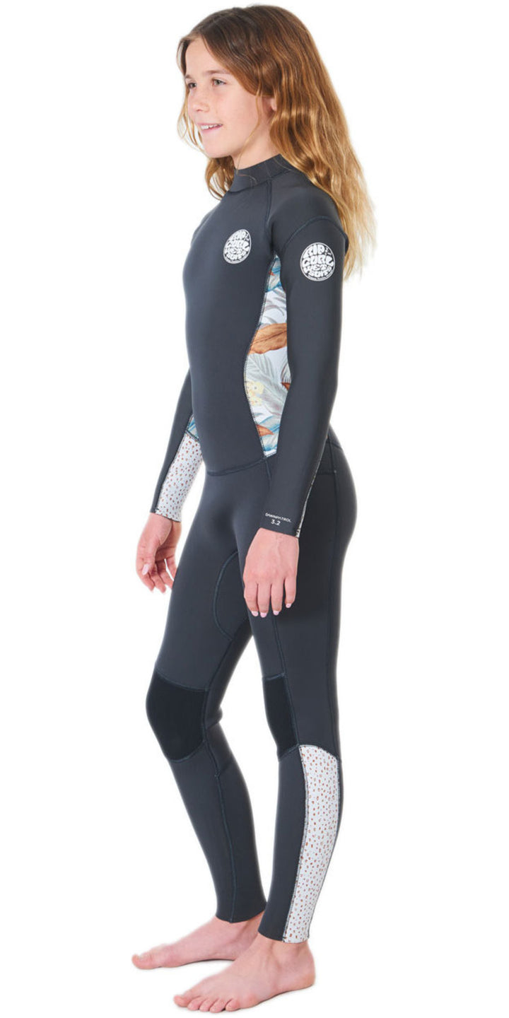 A girl wearing a black and multicolored floral Rip Curl Dawn Patrol 5mm kids wetsuit with a back zip. View of the side